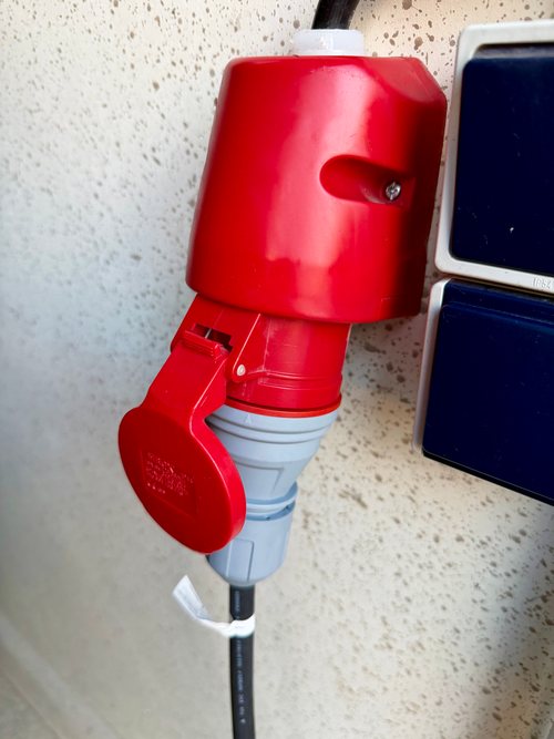 Roter CEE-16-A-Stecker der dé Mobile Ladestation in einer roten CEE-Steckdose an der Hauswand, daneben blaue Außensteckdosen