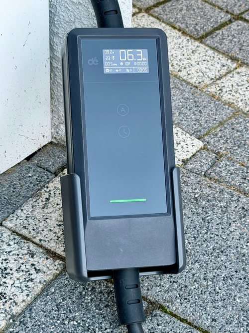 Steuerbox der dé Mobile Ladestation in ihrer Halterung auf Pflastersteinen: Display mit 6,3 kW, darunter Tasten mit A- und Uhrsymbol, unten eine grün leuchtende LED-Leiste
