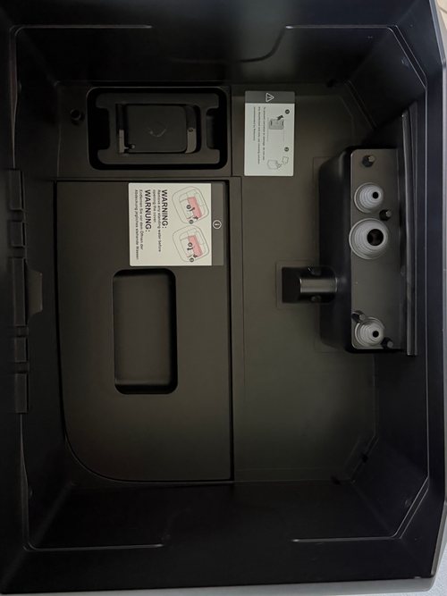 Blick von oben in die Station ohne Wassertanks: links oben das Fach für Reinigungsmittel, darunter der Deckel zum Staubbeutel mit Warnhinweis, rechts die Anschlüsse der Tanks