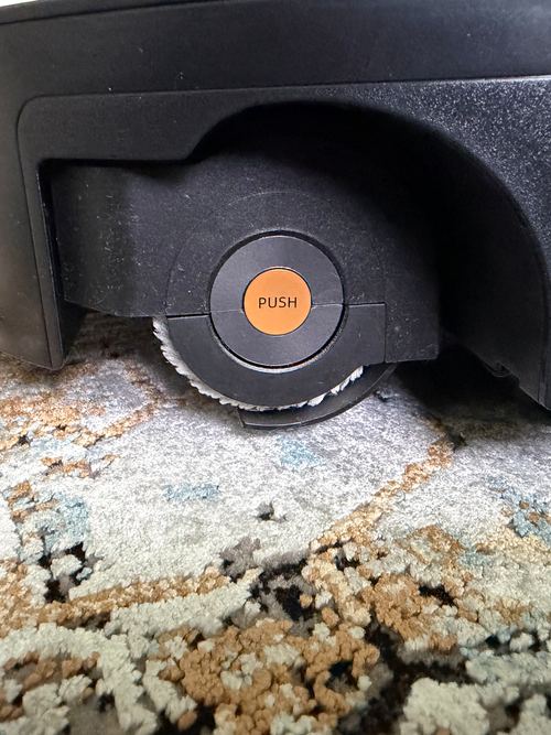 Seitenansicht des Roborock Saros 20 Flow auf einem gemusterten Teppich: Walzenende mit orangefarbener PUSH-Taste, darunter eine schwarze Abdeckung zwischen Walzenbezug und Teppich