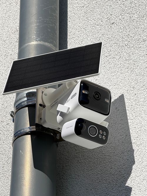 Tapo C675D KIT mit Metallwinkel und zwei Schlauchschellen an einem Fallrohr vor einer weißen Putzfassade, Solarpanel direkt über der Kamera